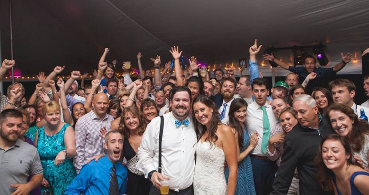 Bill and a huge crowd of wedding guests celebrating with hands raised on the dance floor